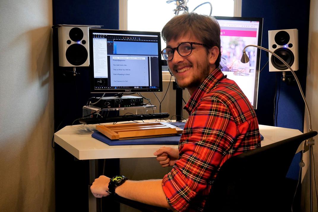 Een voorlezer zit in een studiocabine met beeldscherm en microfoon