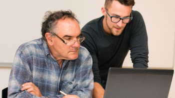 twee mensen kijken naar een beeldscherm; de een zittend, de andere  persoon staat en legt iets uit 